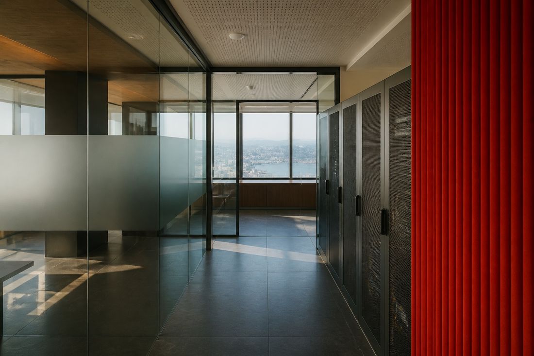 An empty office corridor behind glass partitions, cabinets along one side and a red panel at the end