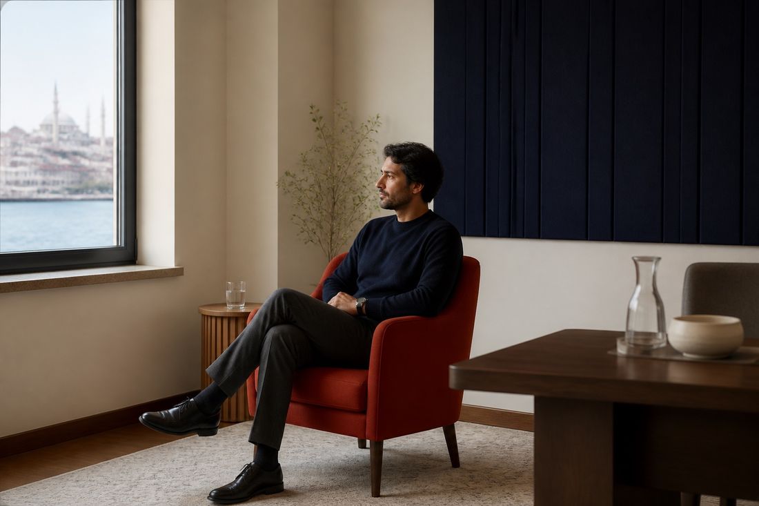 Man in a navy sweater sits alone in a rust-red armchair, hands clasped, looking out a window towards water and a domed mosque