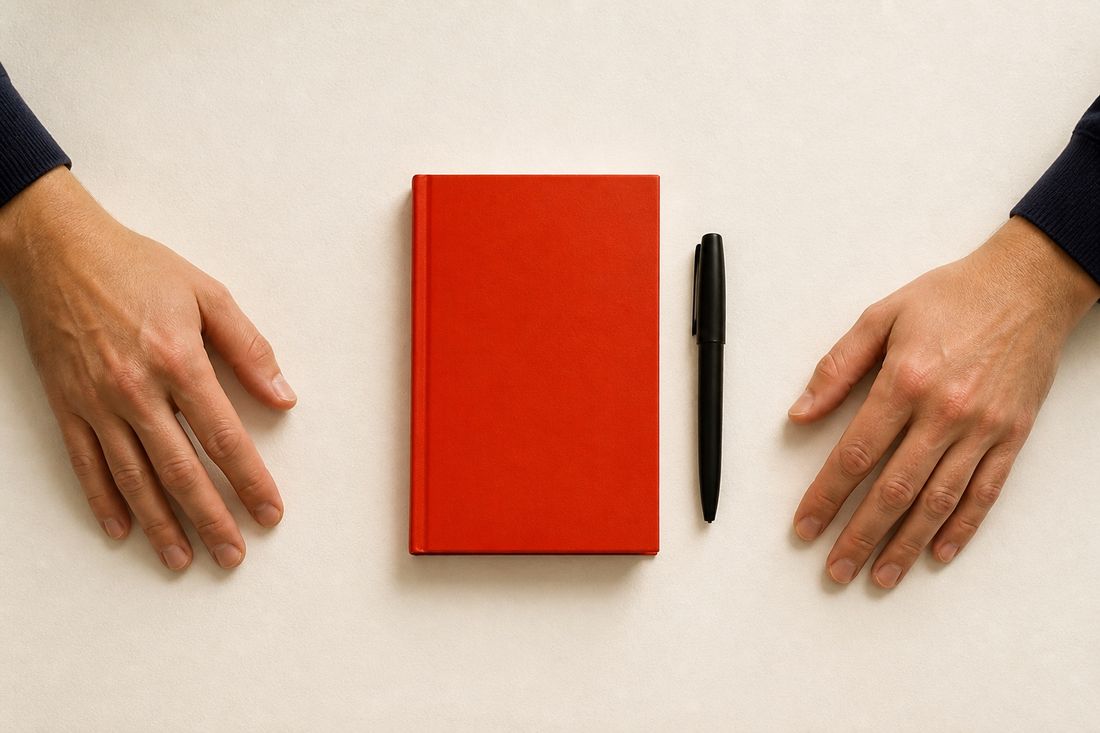 Two hands rest flat on a pale surface either side of a closed red hardback notebook and a black capped pen, navy cuffs showing