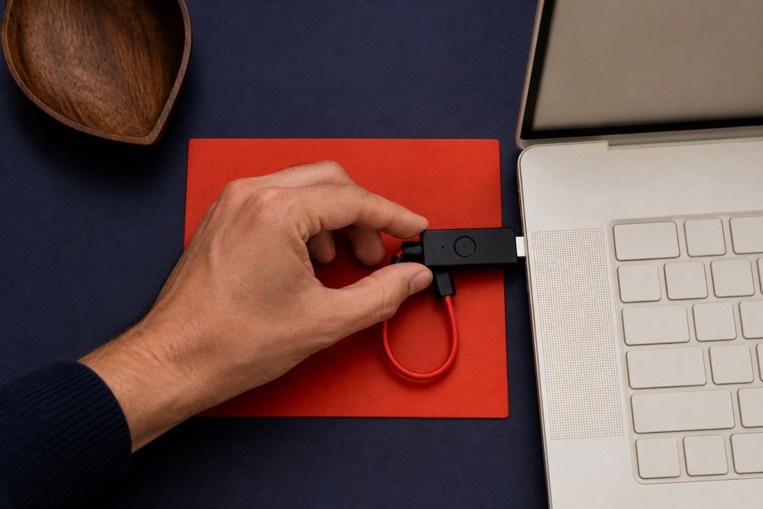 Hand guiding a small black security key on a short red braided cable into a pale laptop, on a red mat over navy felt