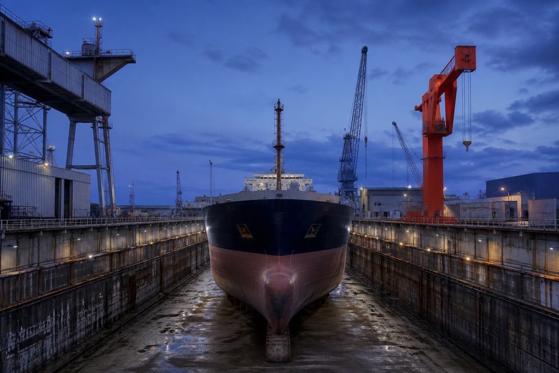 Cargo ship sitting in a drained dry dock at blue dusk, its dark hull and red-brown underside exposed beneath tall shipyard cranes