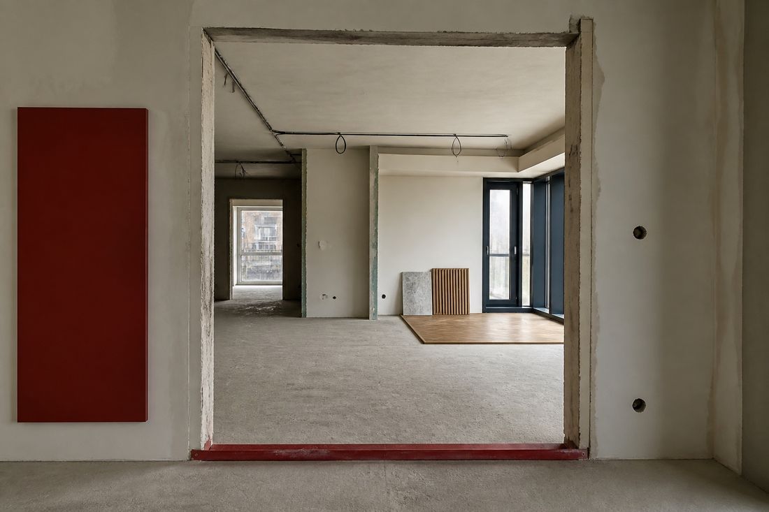 Unfinished apartment: bare screed floors, exposed ceiling conduit, a deep red wall panel, and a patch of new oak flooring