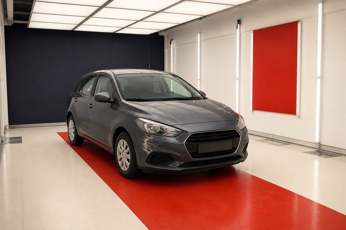 A grey hatchback with a pale scrape above the front wheel arch stands on a red floor mat in a white inspection bay