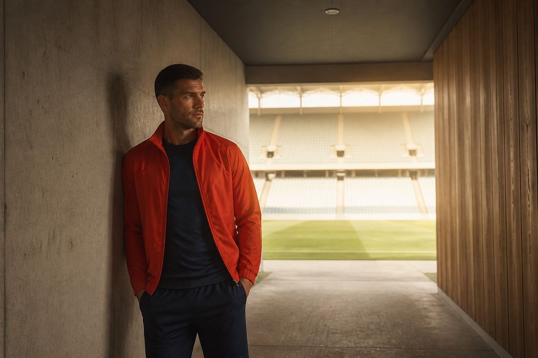 Athlete in a red zip-up track jacket and navy kit leaning against the concrete wall of a stadium tunnel, empty stands beyond